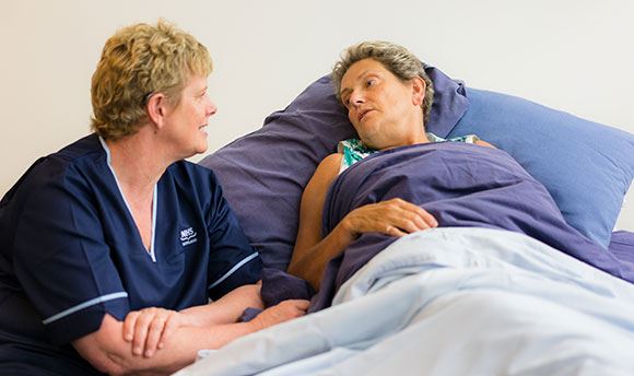 A palliative care nurse sitting by a patient's bed speaking to her