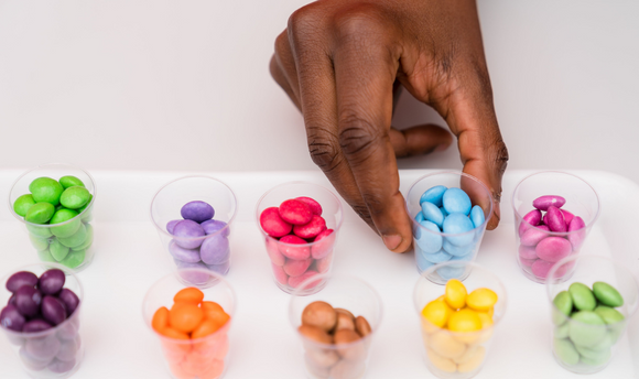 A human hand reaches towards one of 10 tiny containers full of brightly coloured samples of sweets. Each container contains individual colours of sweets.